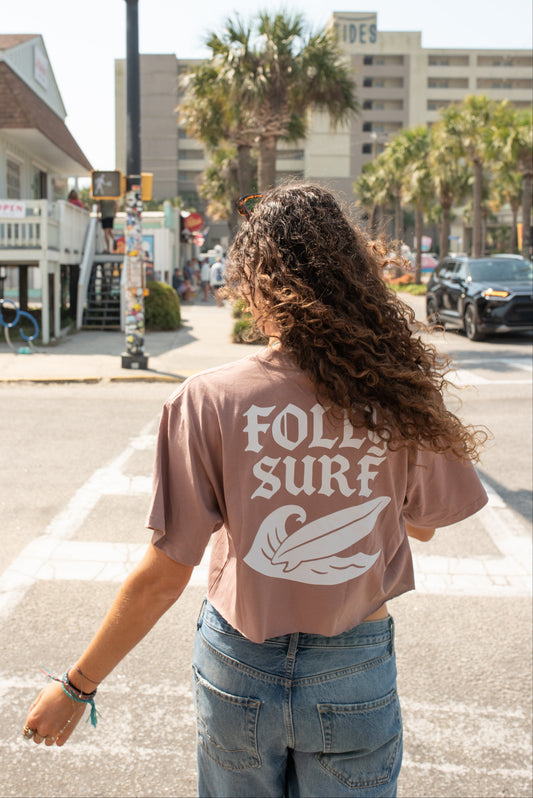 Person wearing a 'Folie Surf' t-shirt walking on a street with palm trees and buildings in the background.
