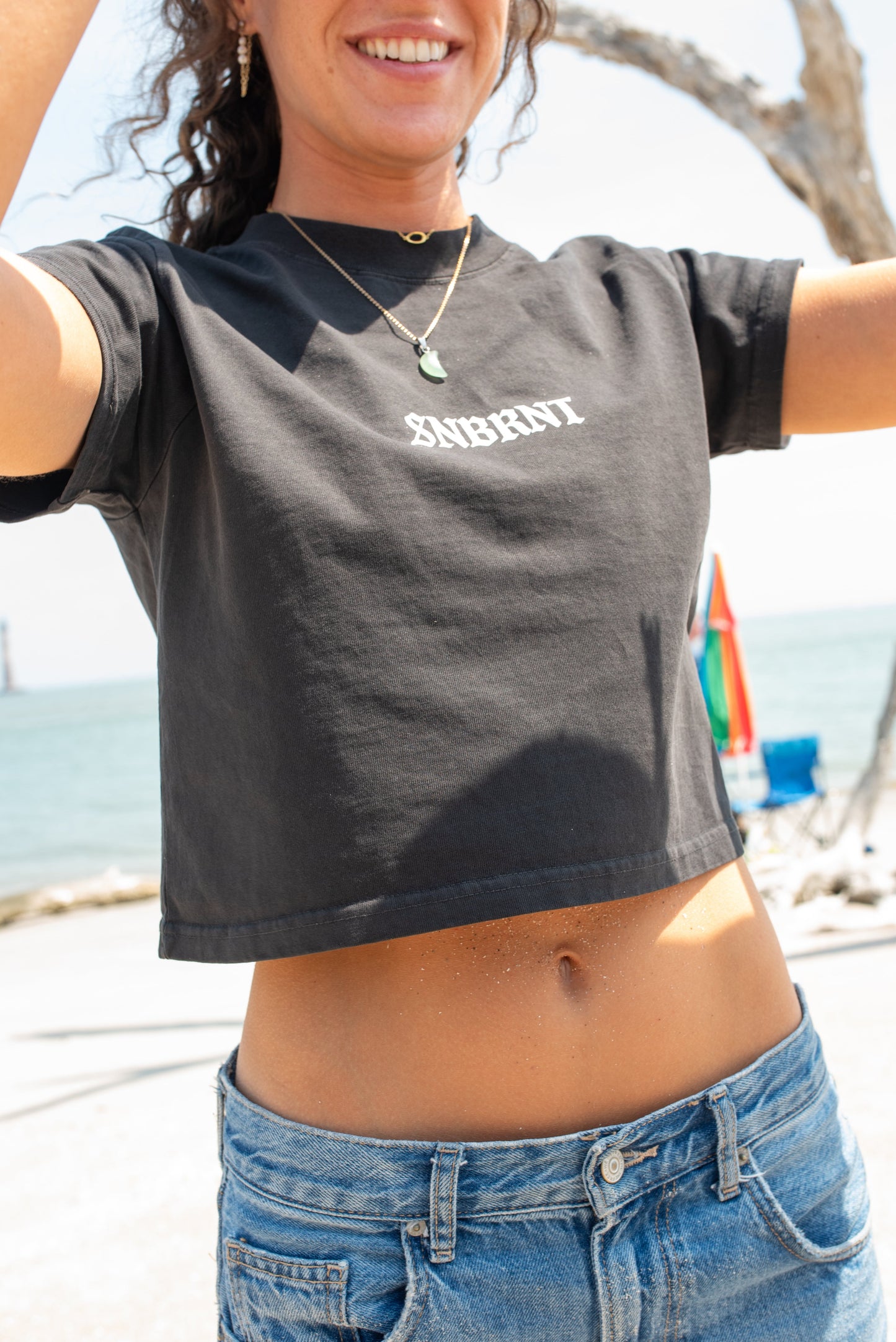 Person wearing a black t-shirt with 'SNBRNT' printed on it at a beach.